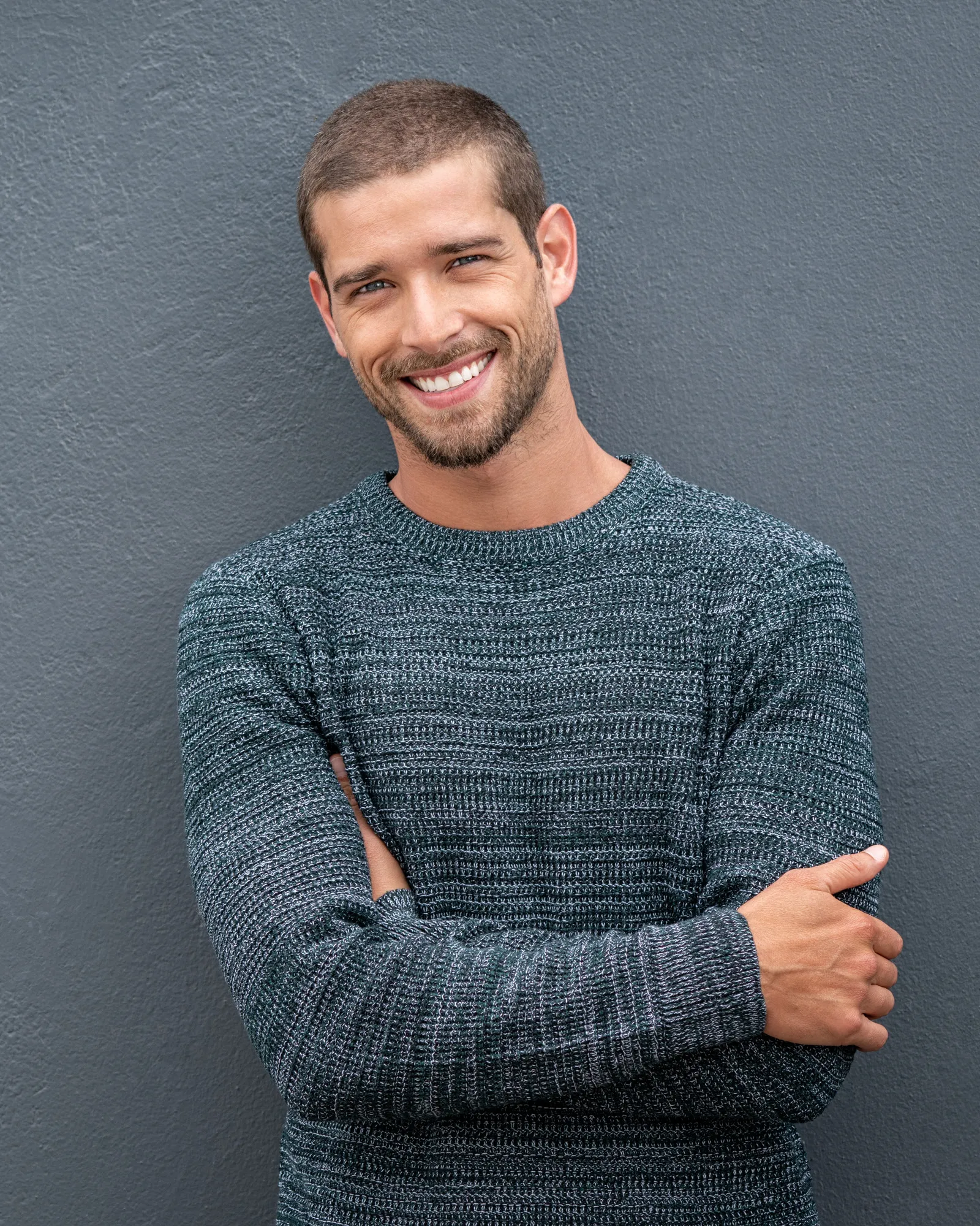 Lächelnder Mann mit kurz geschnittenem Haar und Bart im grauen Strickpullover lehnt mit verschränkten Armen an einer grauen Wand.